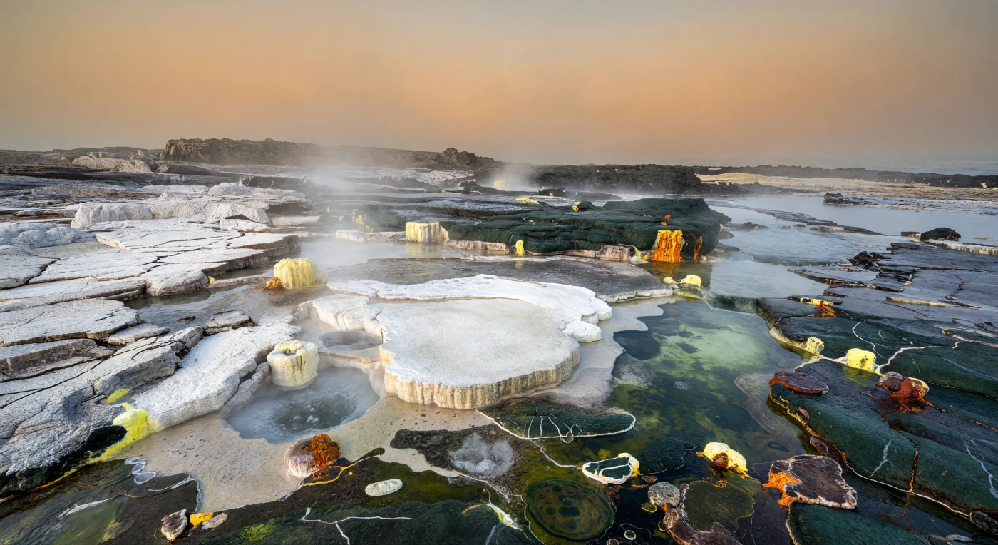 On a low, barren Archean craton margin about 2.9–2.7 billion years ago, silica-rich hot springs and fumaroles spread white sinter, red iron oxides, and yellow sulfur across fractured TTG granitoids and dark greenstone bedrock. Life is visible only as thin green-black to brown-purple microbial films clinging to wet rock beside steaming pools and runoff channels, while all dry surfaces remain starkly lifeless. This scene captures one of Earth’s earliest subaerial hydrothermal landscapes, where microbial communities may have endured on emergent continental crust under an oxygen-poor, haze-filled sky long before plants or animals existed.